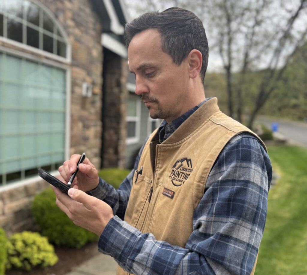 Man in a tan work vest and blue plaid shirt uses a stylus on a smartphone outdoors near a brick building.