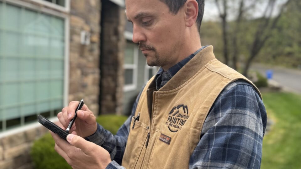 Man in a tan work vest and blue plaid shirt uses a stylus on a smartphone outdoors near a brick building.