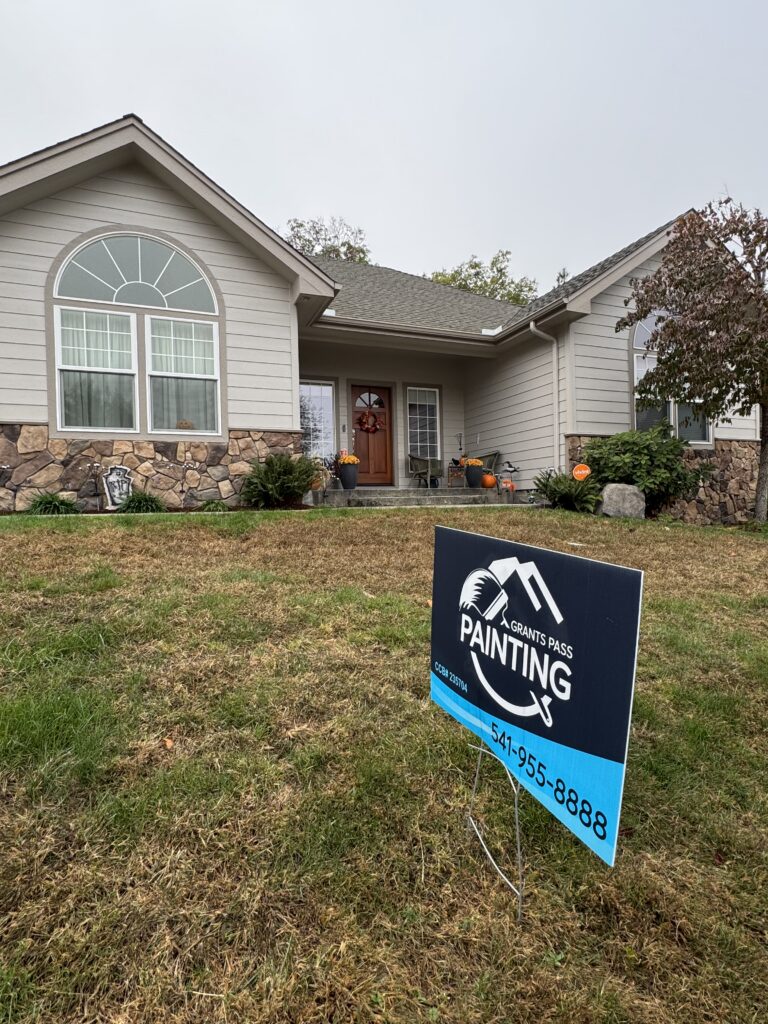 Front view of a beige house with stone foundation and fall decorations by the entry, plus a blue painting sign in the yard.