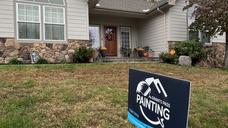 Front view of a beige house with stone foundation and fall decorations by the entry, plus a blue painting sign in the yard.