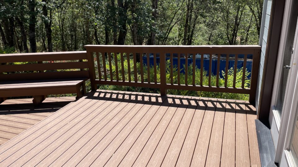 Wooden deck with railing, facing a dense stand of trees; a blue container is visible beyond the railing behind the yard.