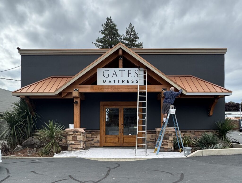 Worker on a ladder hanging a sign that reads 'GATES MATTRESS' on a storefront with dark exterior and wooden trim.