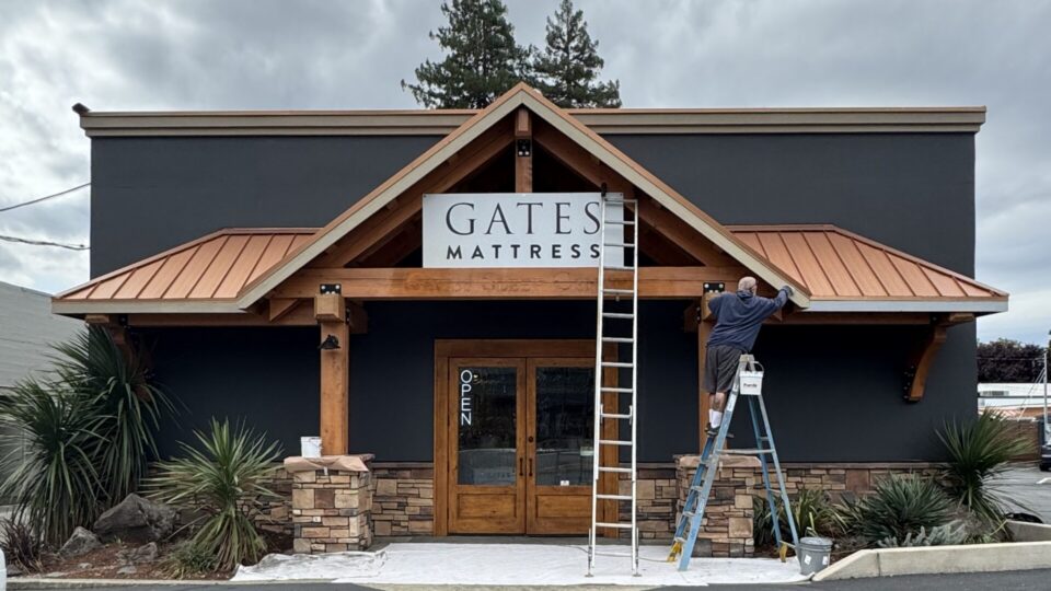 Worker on a ladder hanging a sign that reads 'GATES MATTRESS' on a storefront with dark exterior and wooden trim.