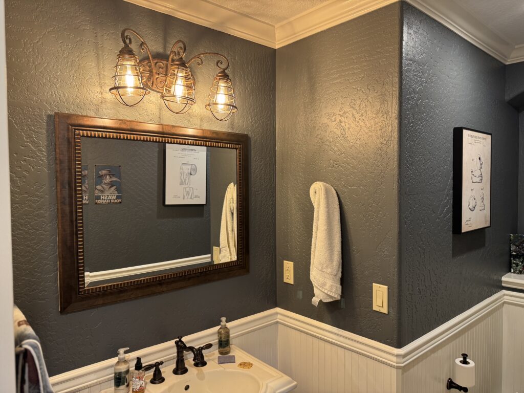Bathroom vanity area with a bronze-framed mirror, three industrial cage lights, and dark textured walls.