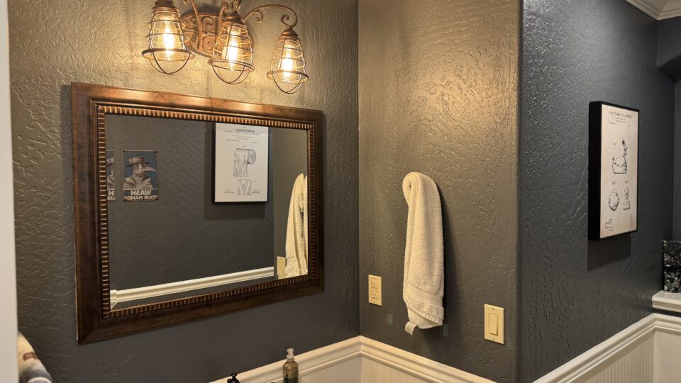 Bathroom vanity area with a bronze-framed mirror, three industrial cage lights, and dark textured walls.
